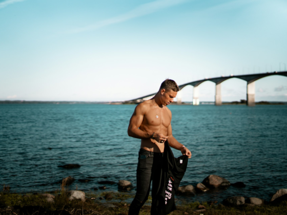 A shirtless ripped guy standing in front of a bridged river in an aesthetic manner
