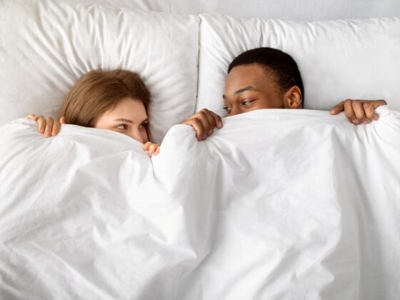 couple hiding under white blanket in bed, looking at each other with affection